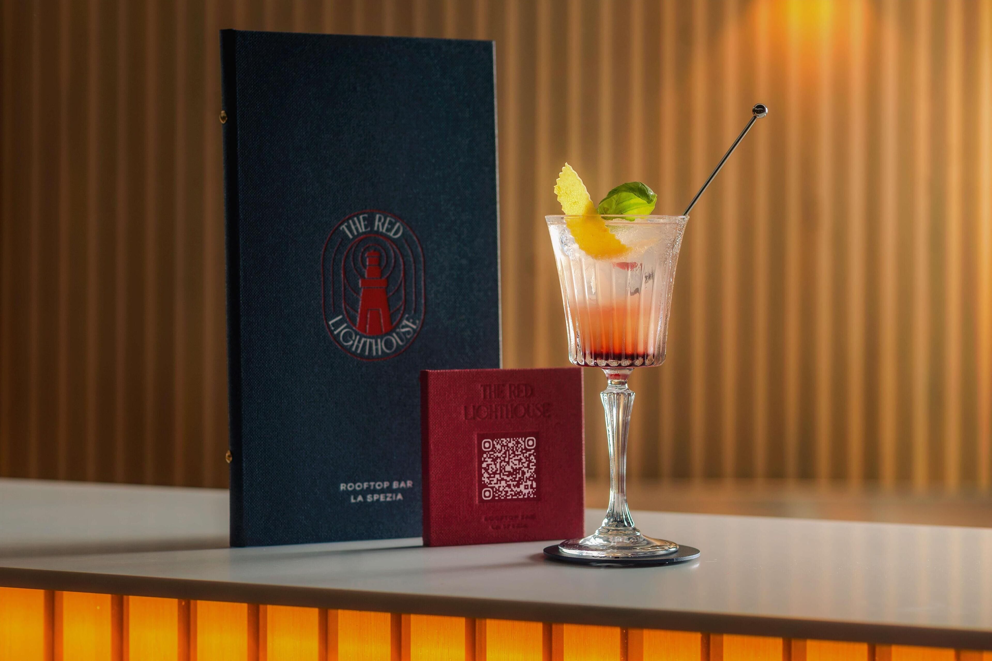 A cocktail glass with a lemon twist garnish on a bar counter, a QR code, and a menu titled "The Red Lighthouse" next to it.