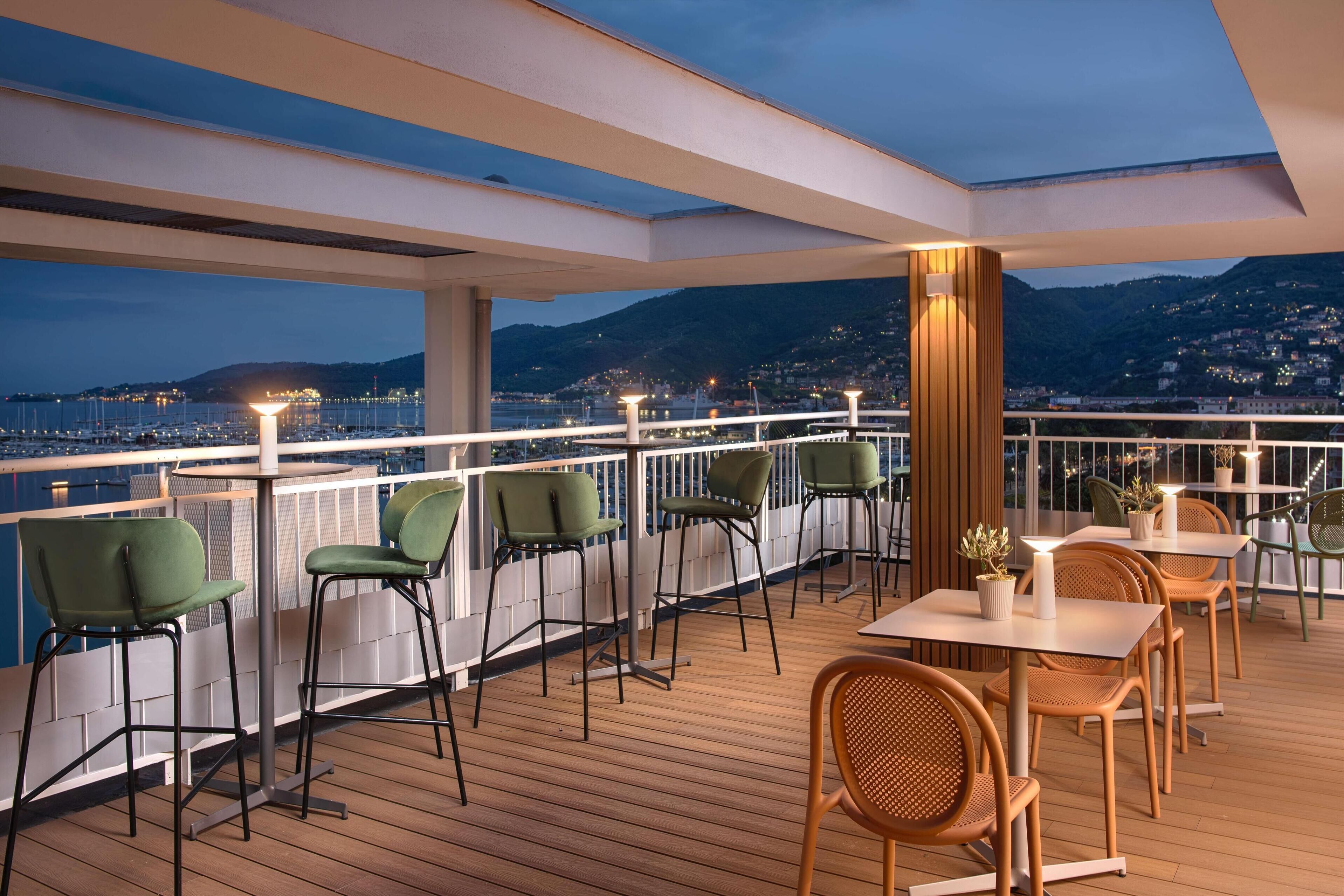 A deck with wooden flooring, green and wooden chairs, and tables with potted plants.