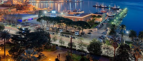 Un paisaje urbano costero al atardecer con un puerto deportivo, instalaciones portuarias y una carretera bordeada de coches aparcados y palmeras.