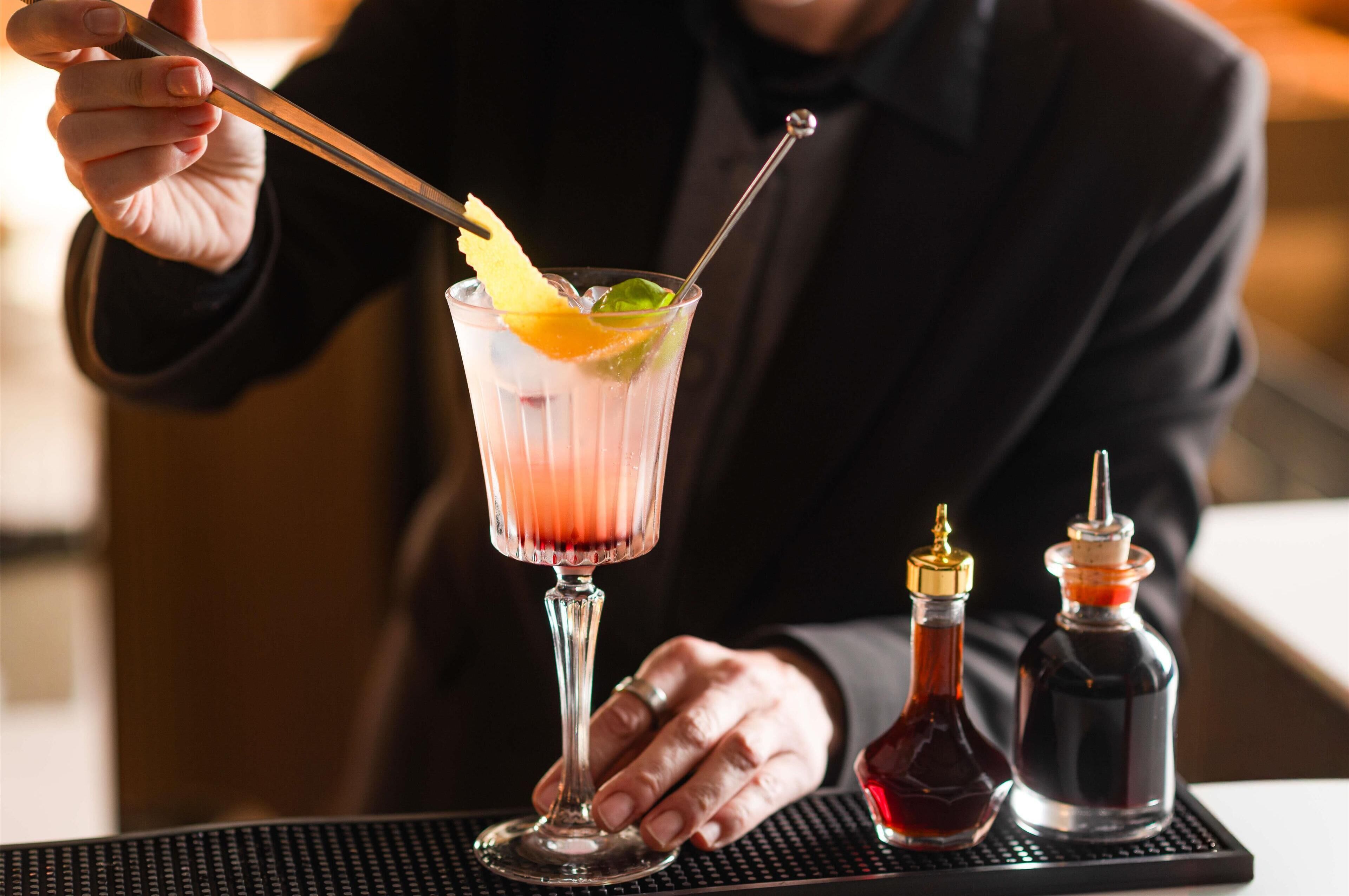 En bartender som blandar en cocktail med hjälp av en shaker, ett glas och två flaskor sås, allt placerat på en barbricka.