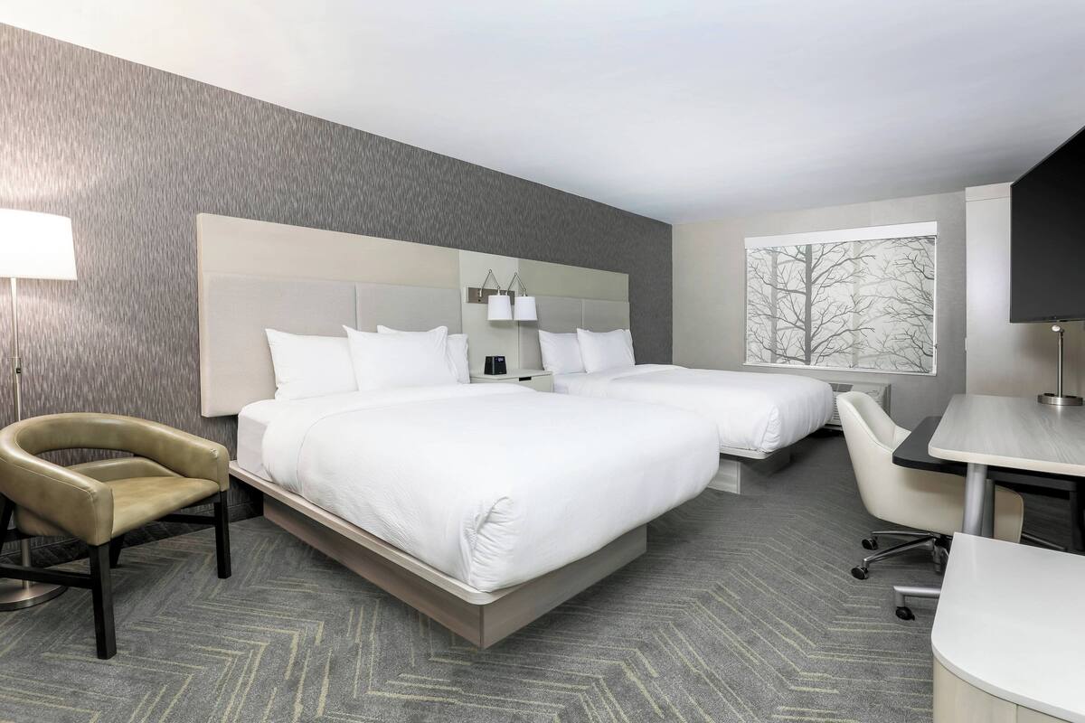 A hotel room with two beds, a desk, a chair, and a large window with a view of trees.