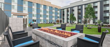 Un patio al aire libre con un elemento decorativo de fuego, mobiliario de mimbre y un edificio moderno al fondo.