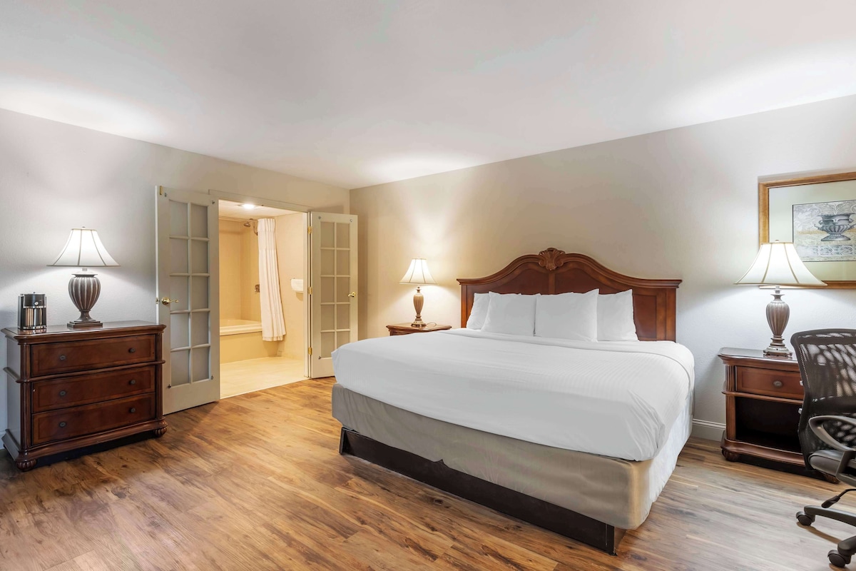Habitación de hotel con una cama grande, mesitas de noche de madera, un escritorio con una silla y una puerta que da a un baño.