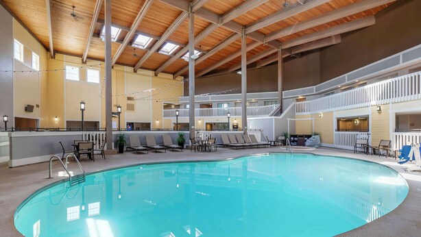 An indoor swimming pool with a wooden ceiling, multiple levels, and lounge areas with chairs and tables.