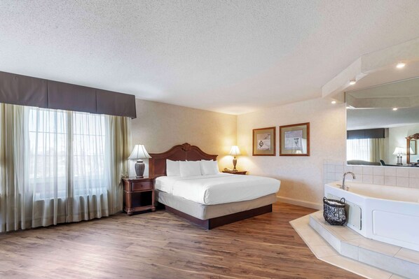 A hotel room with a wooden floor, a bed with white linens, a nightstand with a lamp, a bathroom with a bathtub, and a large window with sheer curtains.