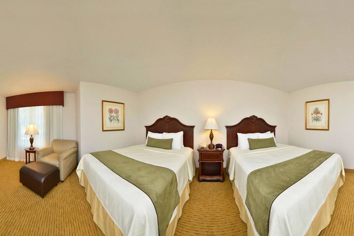 A hotel room with two beds, a chair, a nightstand, a lamp, and framed artwork on the walls.