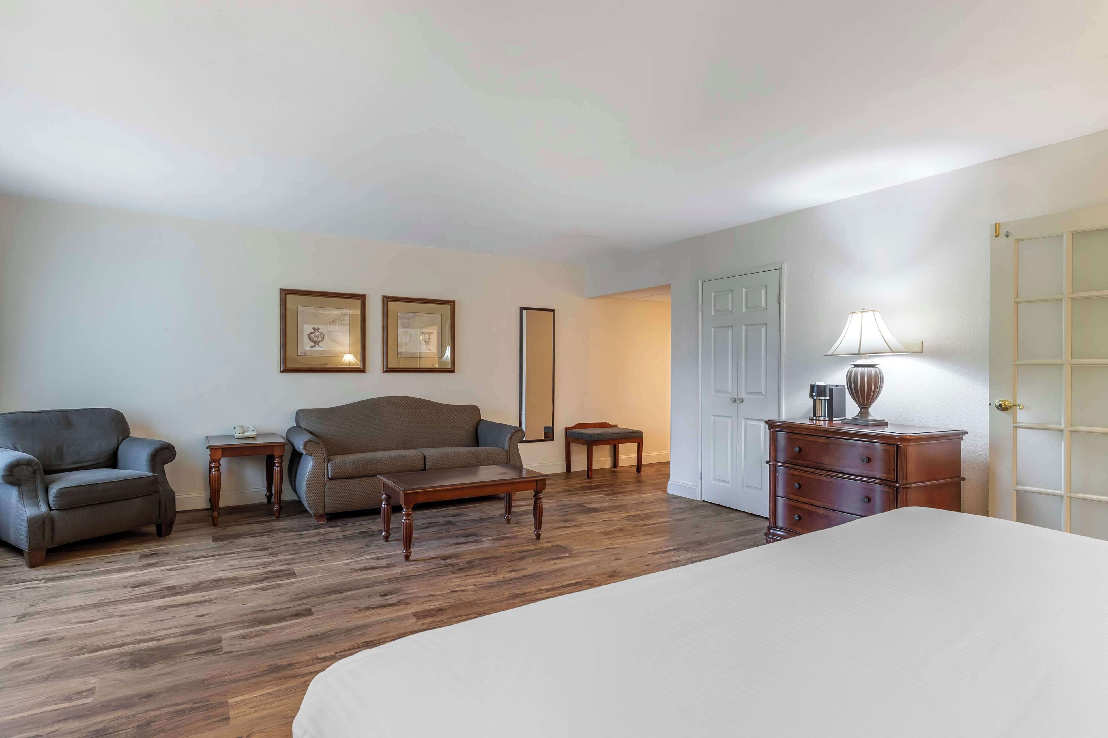 A hotel room with a large bed, a sofa, two armchairs, a wooden dresser, a nightstand, and a door with a frosted glass panel.