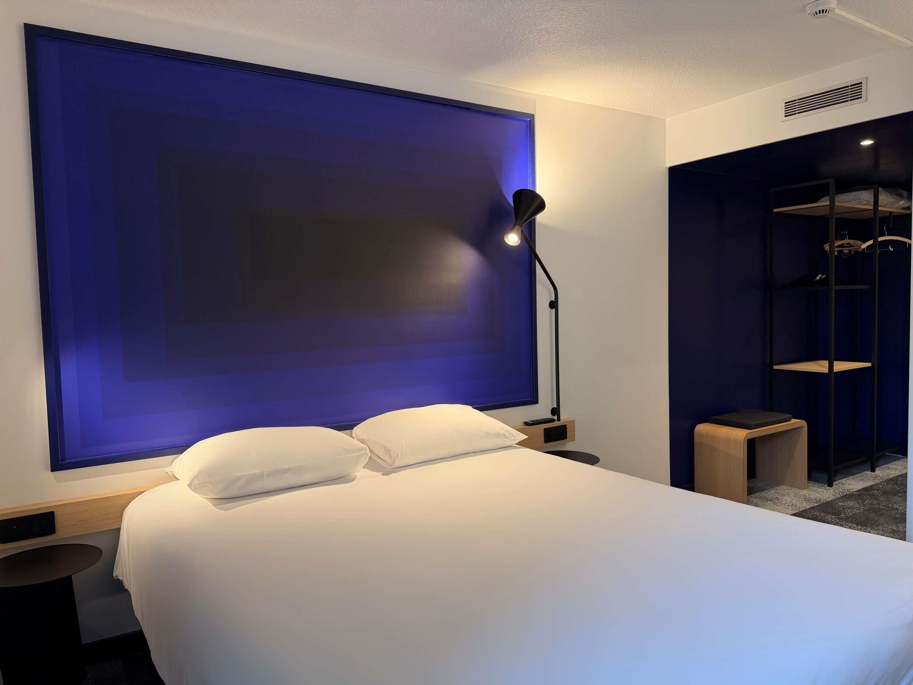 Una habitación de hotel moderna con una cama grande, una mesita de noche, una lámpara y un armario con estantes.