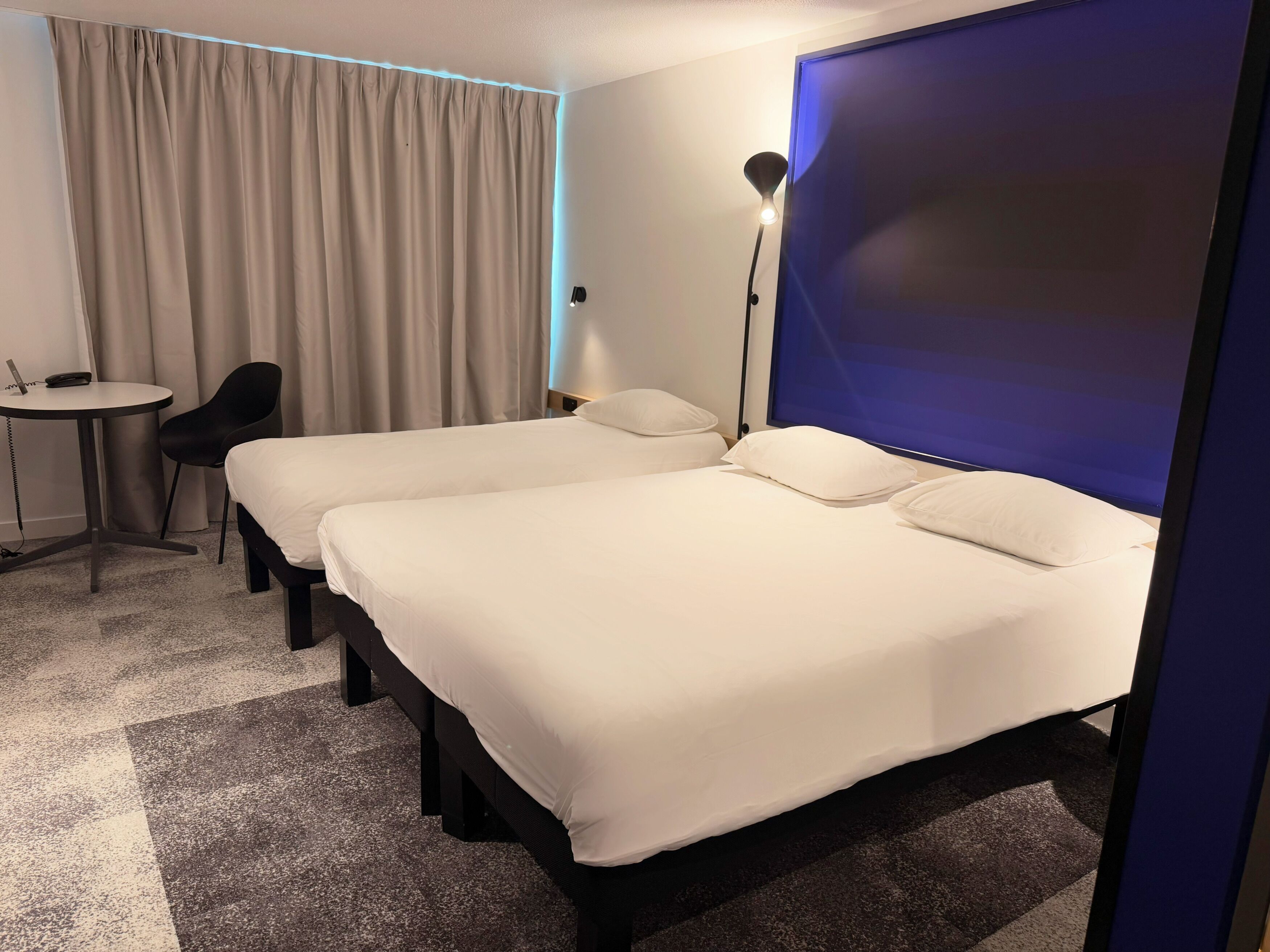 Habitación de hotel con dos camas, un escritorio, una silla, una lámpara y una cortina.
