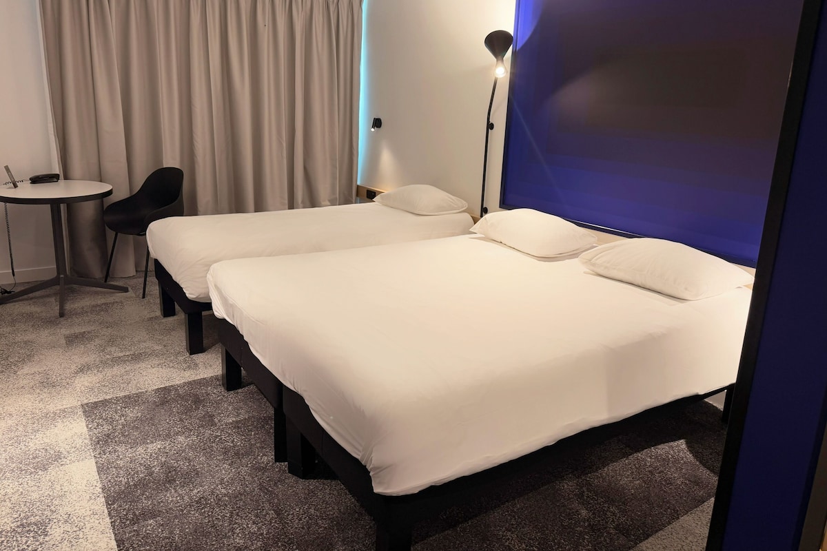 Habitación de hotel con dos camas, un escritorio, una silla, una lámpara y una cortina.