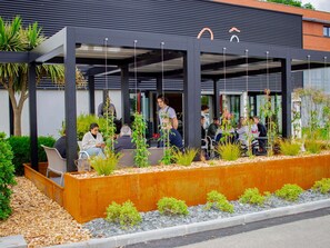 Un espacio al aire libre con mesas y sillas, rodeado de plantas y un muro bajo de color naranja.