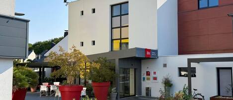 A modern building with a flat roof, large windows, and a sign for 'ibis' indicating a hotel. There are outdoor seating areas with red planters and a bicycle parked nearby.