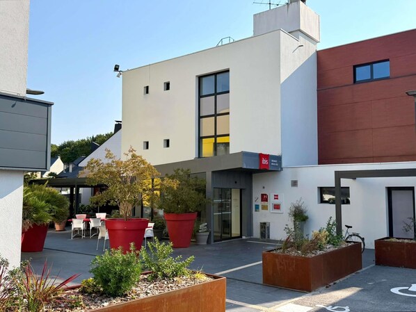 A modern building with a flat roof, large windows, and a sign for 'ibis' indicating a hotel. There are outdoor seating areas with red planters and a bicycle parked nearby.
