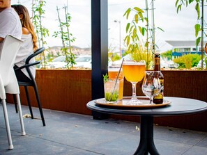Una mesa con bebidas y una botella, una persona sentada en una silla, y plantas al fondo.