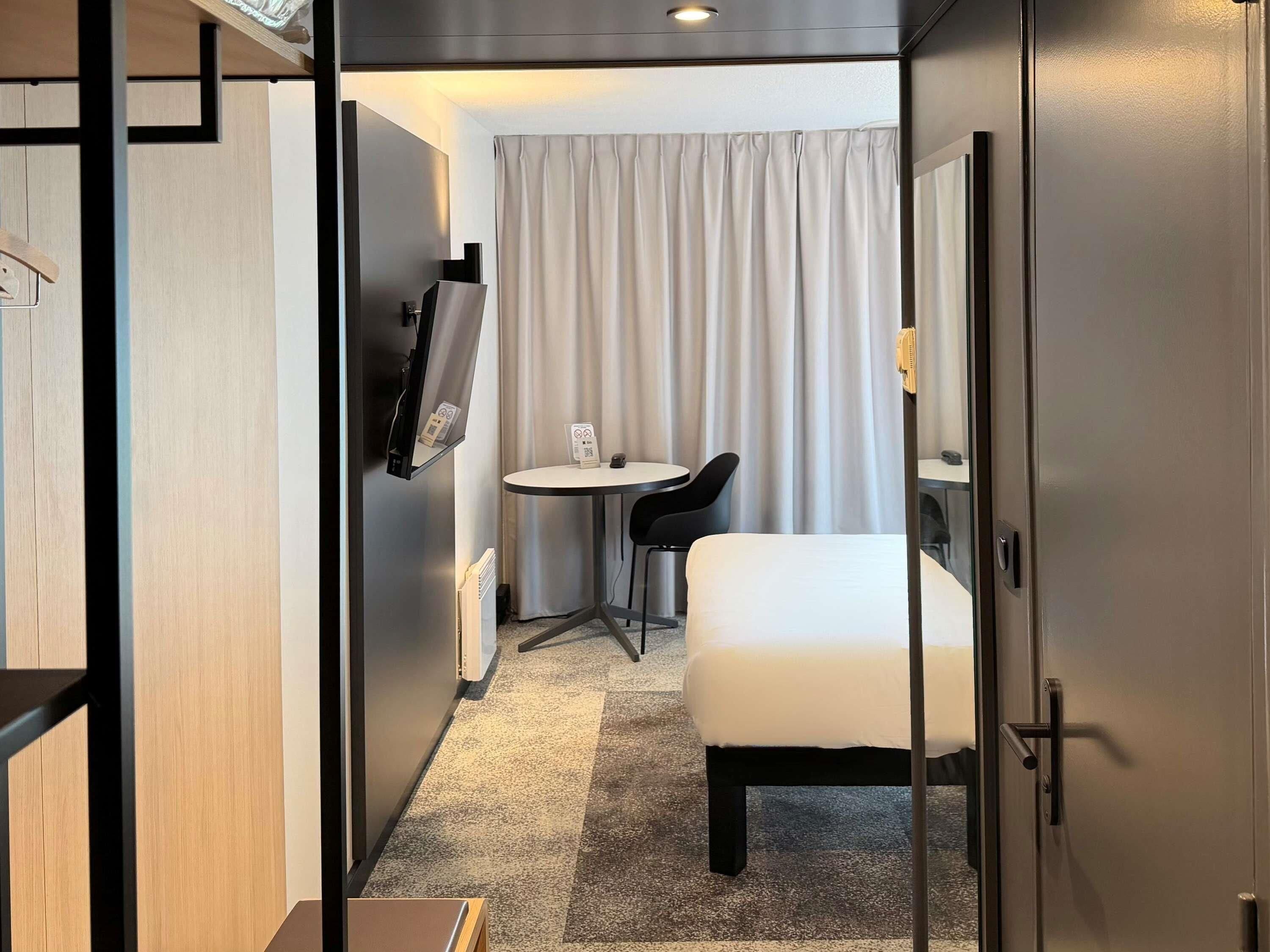 Habitación de hotel con cama, escritorio, silla, televisor y ventana con cortinas.