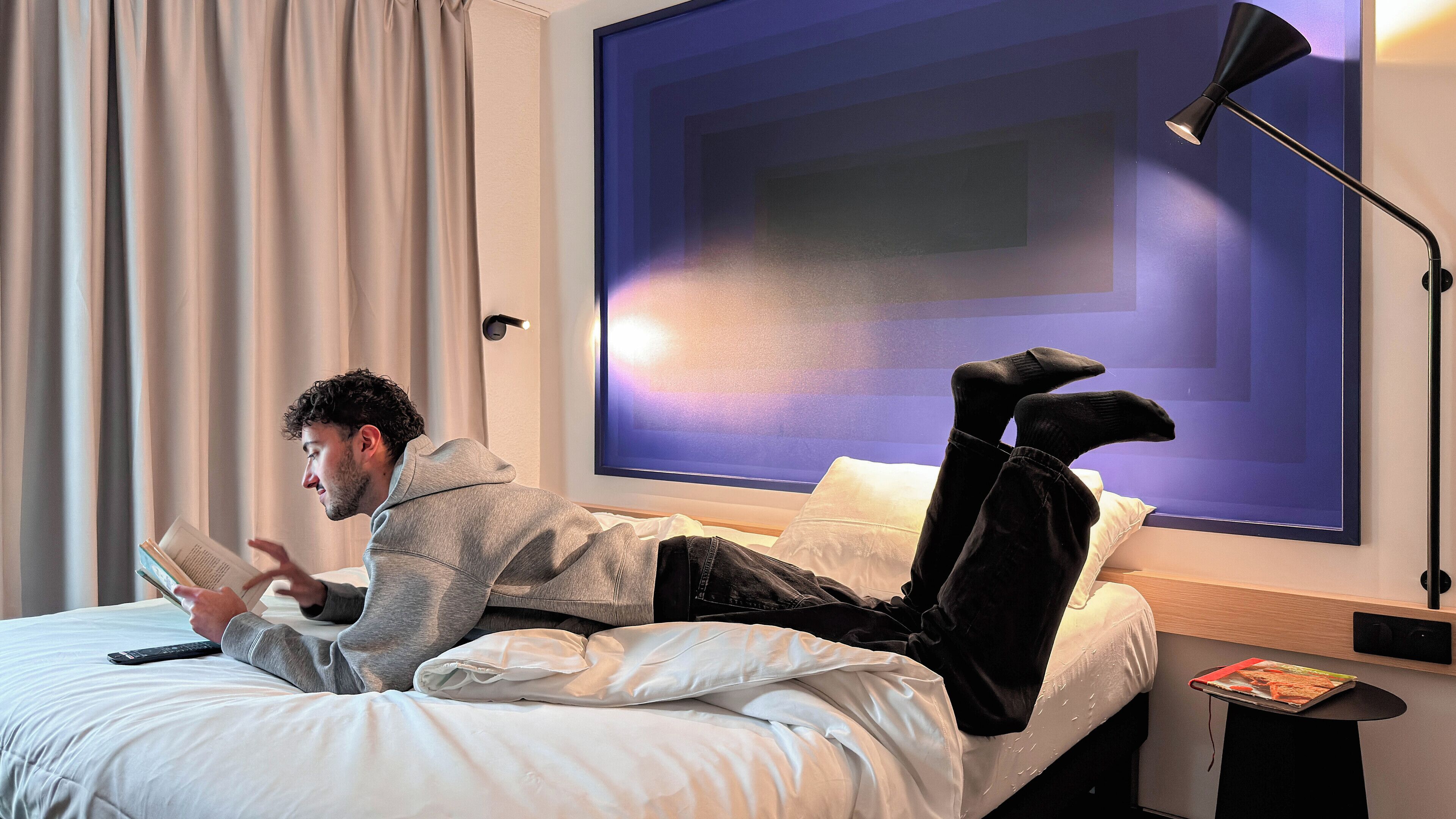 A person lying on a bed in a room with a large window, reading a book.
