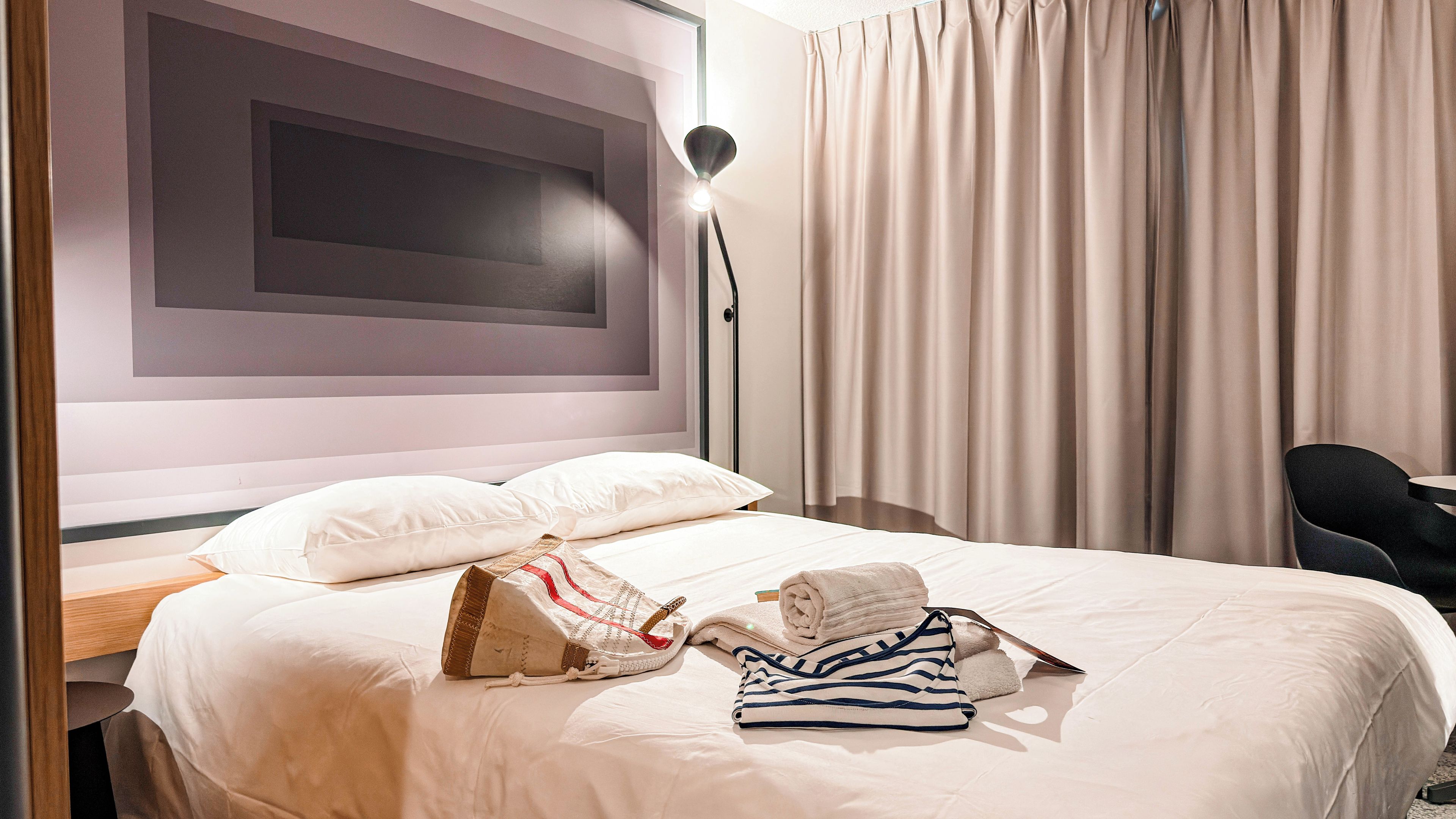 A neatly made bed with pillows, a folded towel, and a handbag on a hotel room with a large window, a floor lamp, and a chair.