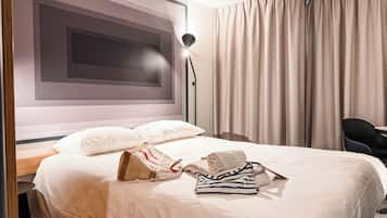 A neatly made bed with pillows, a folded towel, and a handbag on a hotel room with a large window, a floor lamp, and a chair.