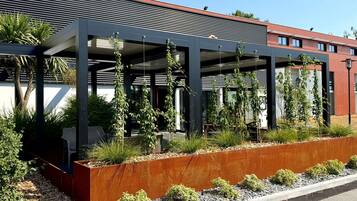 Un bâtiment moderne doté d’un espace extérieur couvert, entouré de verdure et délimité par une clôture en métal.