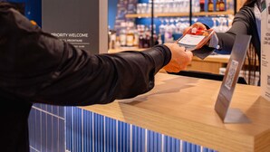 A person using a mobile device at a reception desk in a hotel lobby with a blue and white striped wall and shelves of bottles in the background. - ibis Rennes Beaulieu (Cesson-Sevigne)