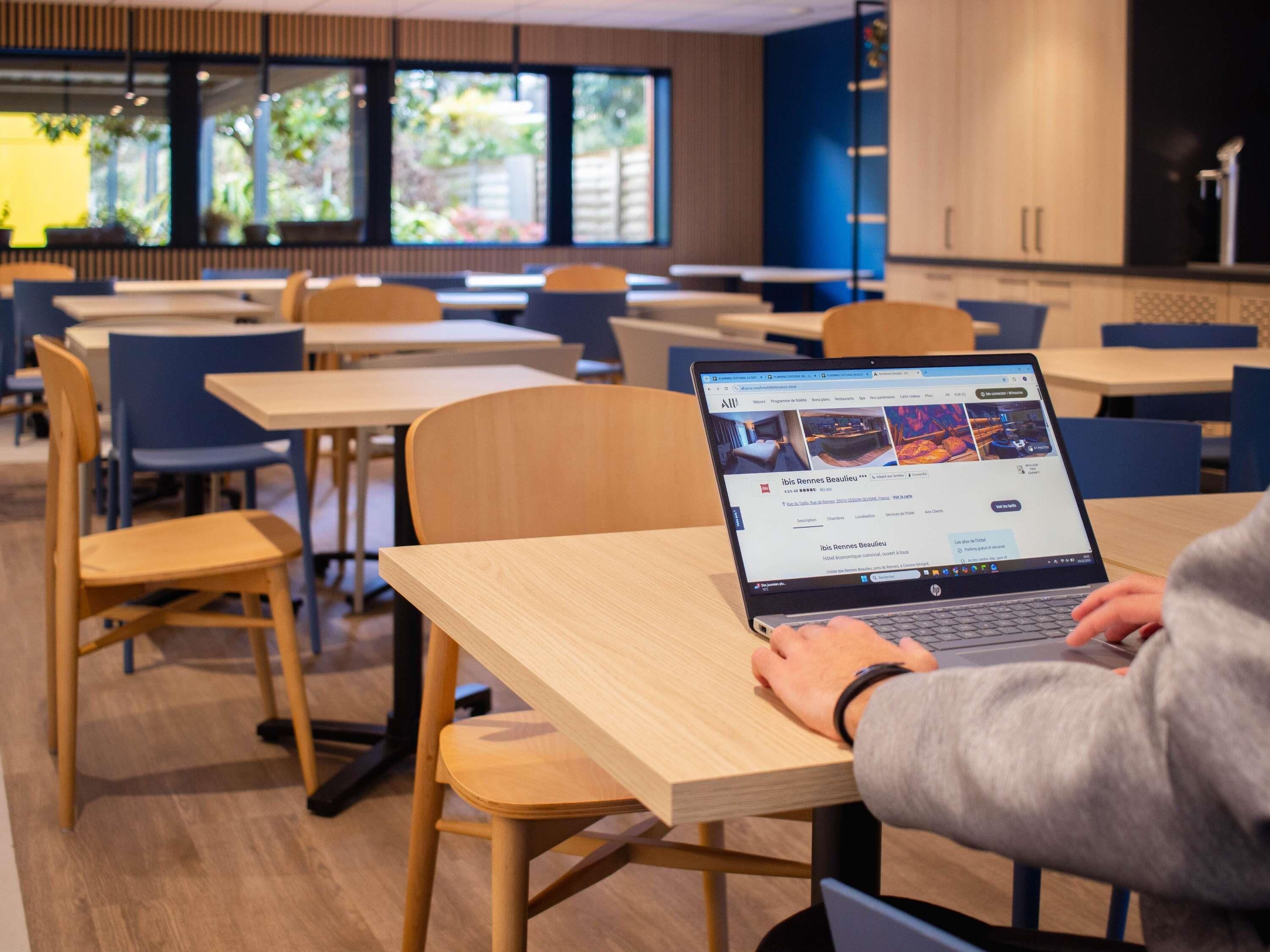 A person using a laptop in a modern, well-lit room with wooden furniture and large windows.