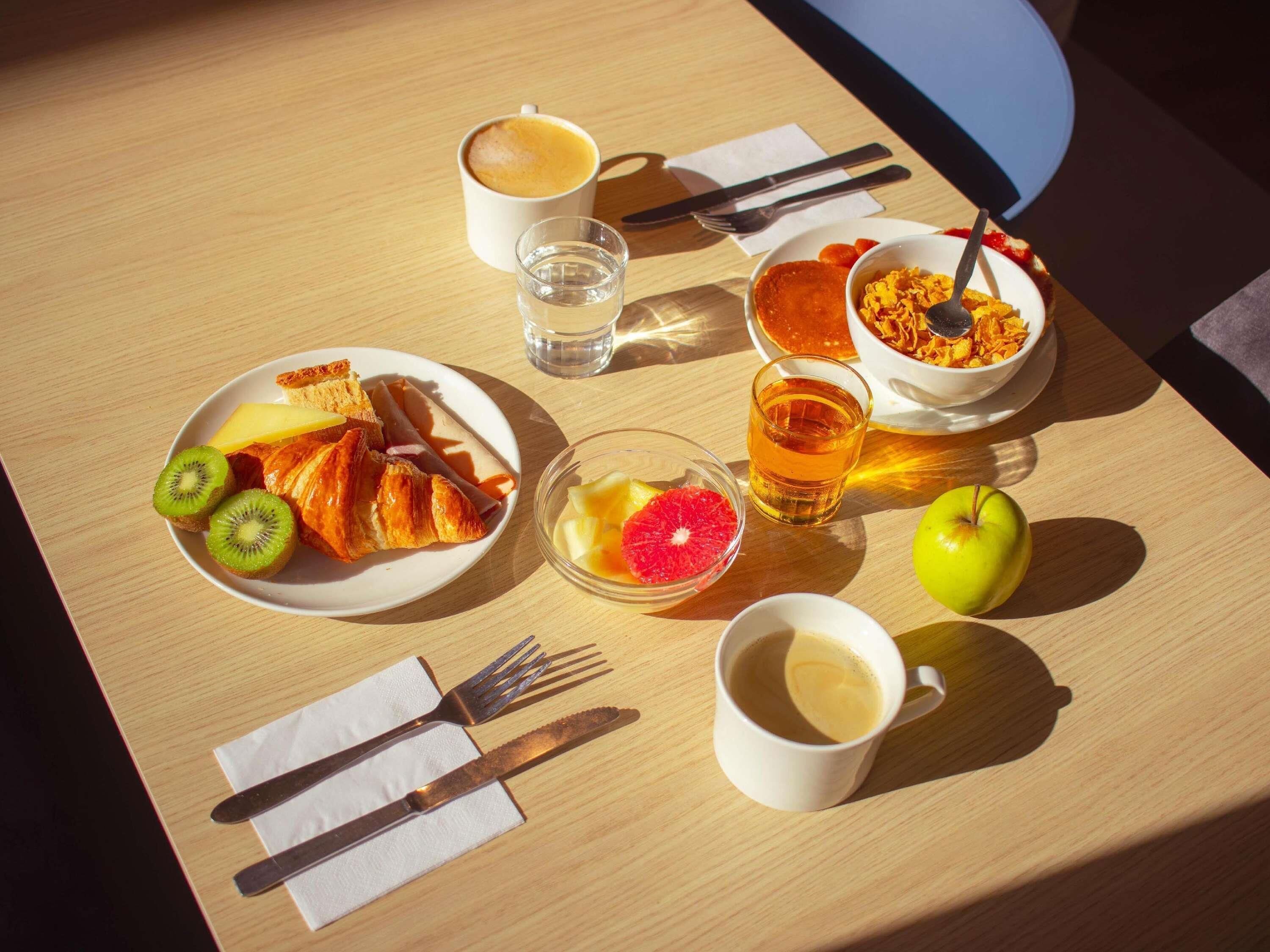 A breakfast table with croissants, kiwi, orange, a glass of juice, a bowl of cereal, a cup of coffee, and a glass of water.