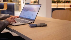 A laptop on a table with a smartphone beside it, in a modern indoor setting with seating and plants in the background. - ibis Rennes Beaulieu (Cesson-Sevigne)