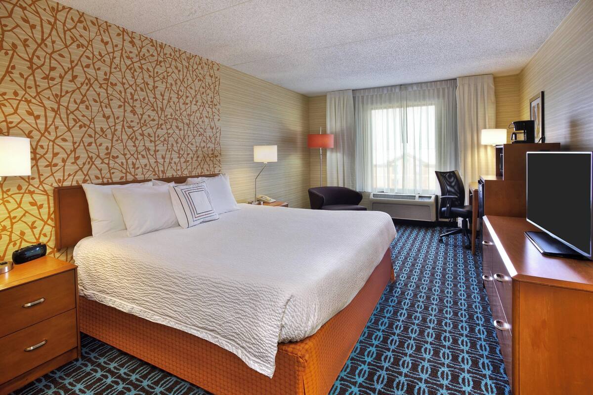 A hotel room with a large bed, a desk with a flat-screen TV, a chair, a desk lamp, and a patterned carpet.