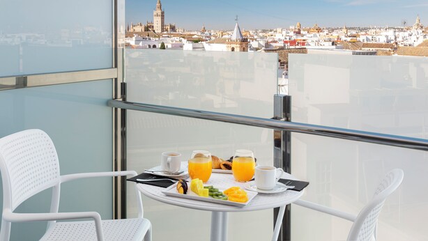 A balcony with a table set for breakfast, including croissants, fruit, and orange juice, overlooking a cityscape with historic buildings.