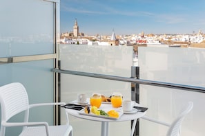Un balcón con una mesa dispuesta para el desayuno, incluyendo cruasanes, fruta y zumo de naranja, con vistas a un paisaje urbano con edificios históricos.