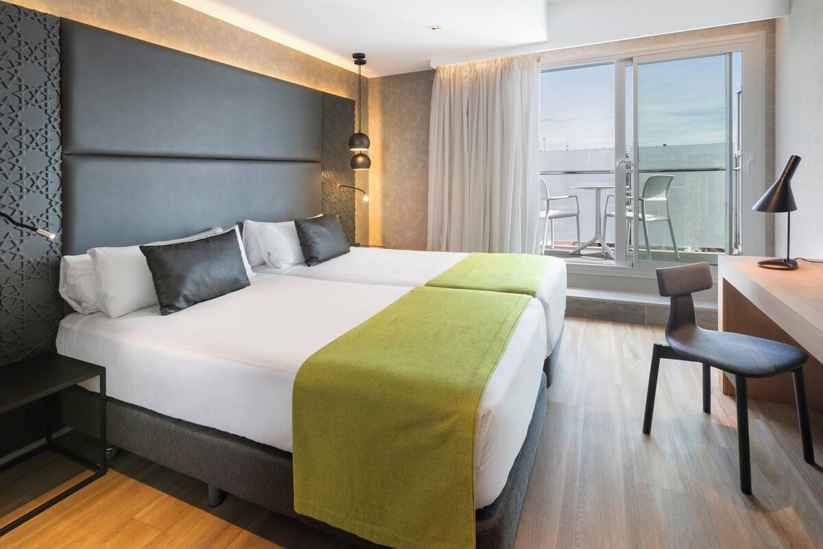 A hotel room with two beds, a desk, a chair, and a balcony with a view.