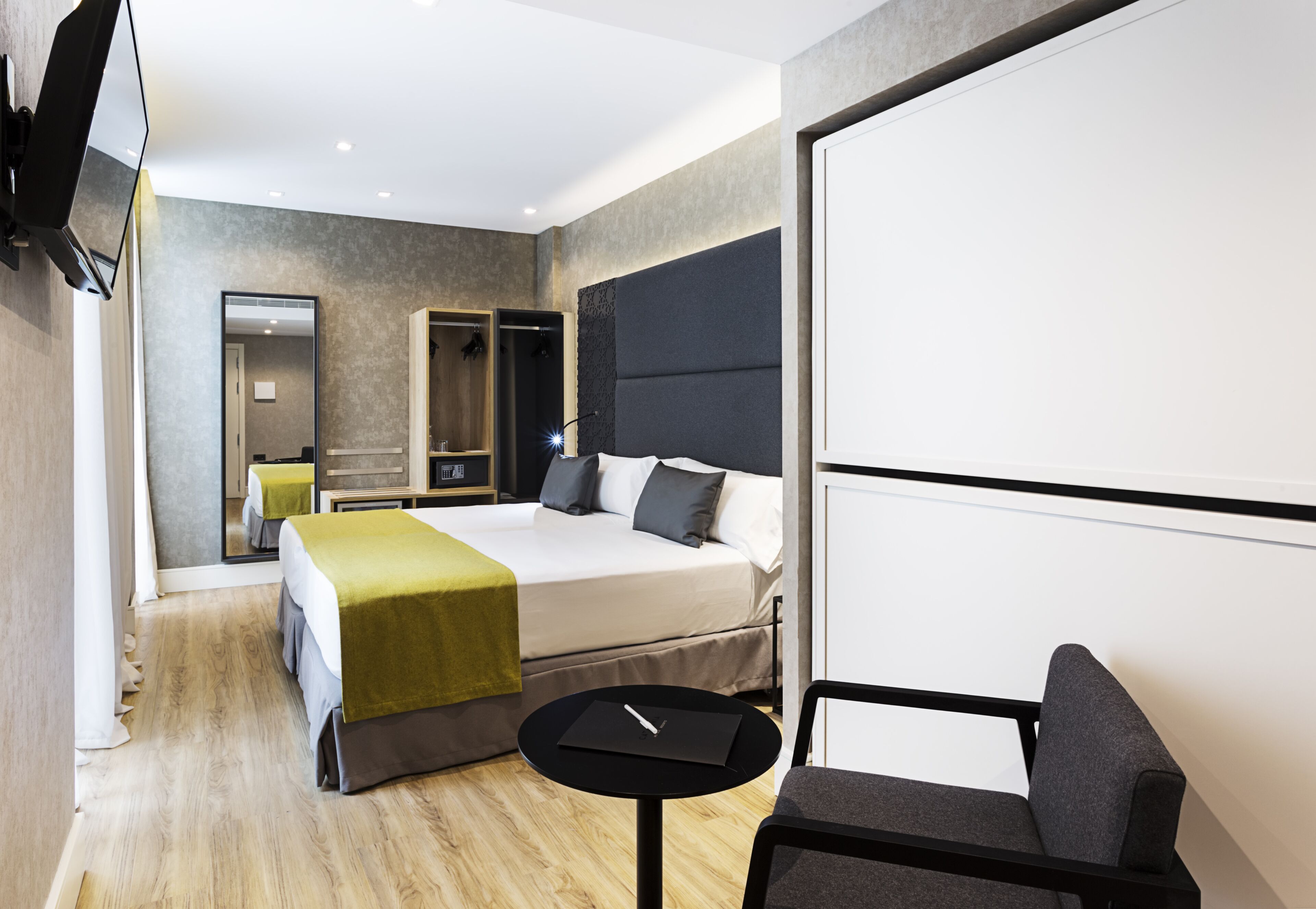 A modern hotel room with a bed, a small table, a chair, a TV, and a mirror.