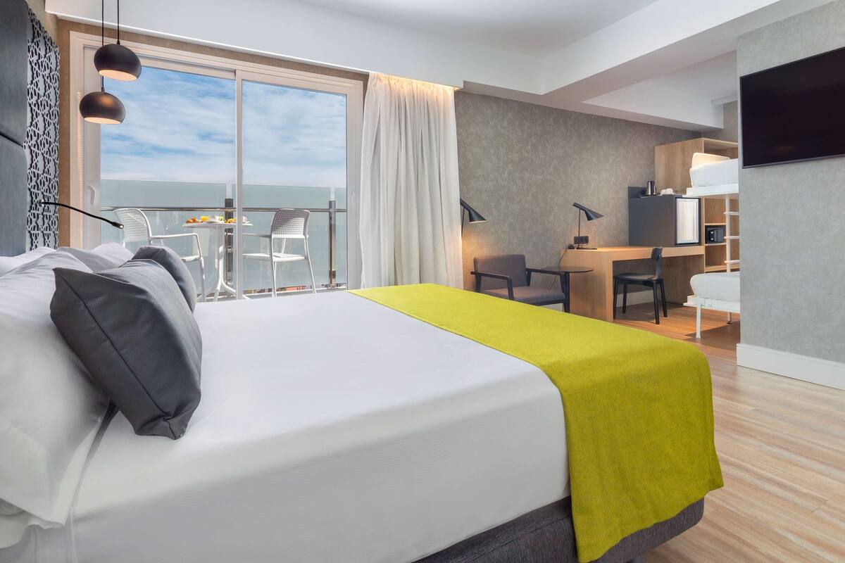 A modern hotel room with a large bed, a balcony with a table and chairs, a desk with a chair, and a TV mounted on the wall.
