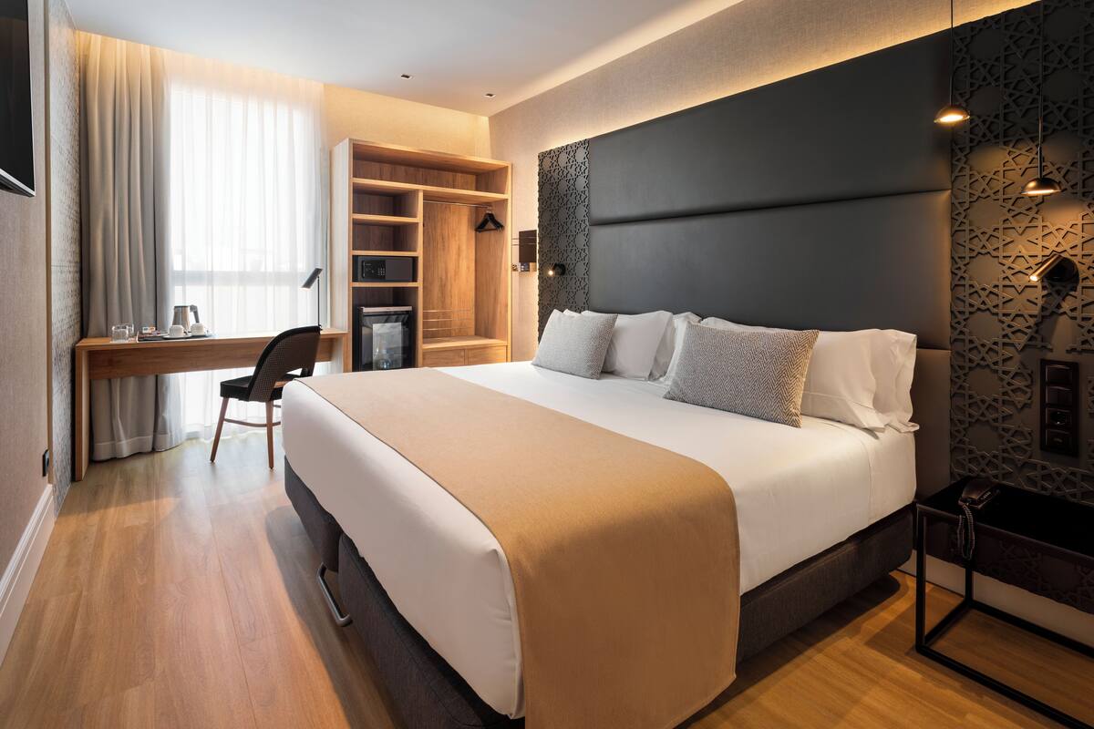 A modern hotel room with a large bed, a desk with a chair, a wardrobe, and a patterned wall.