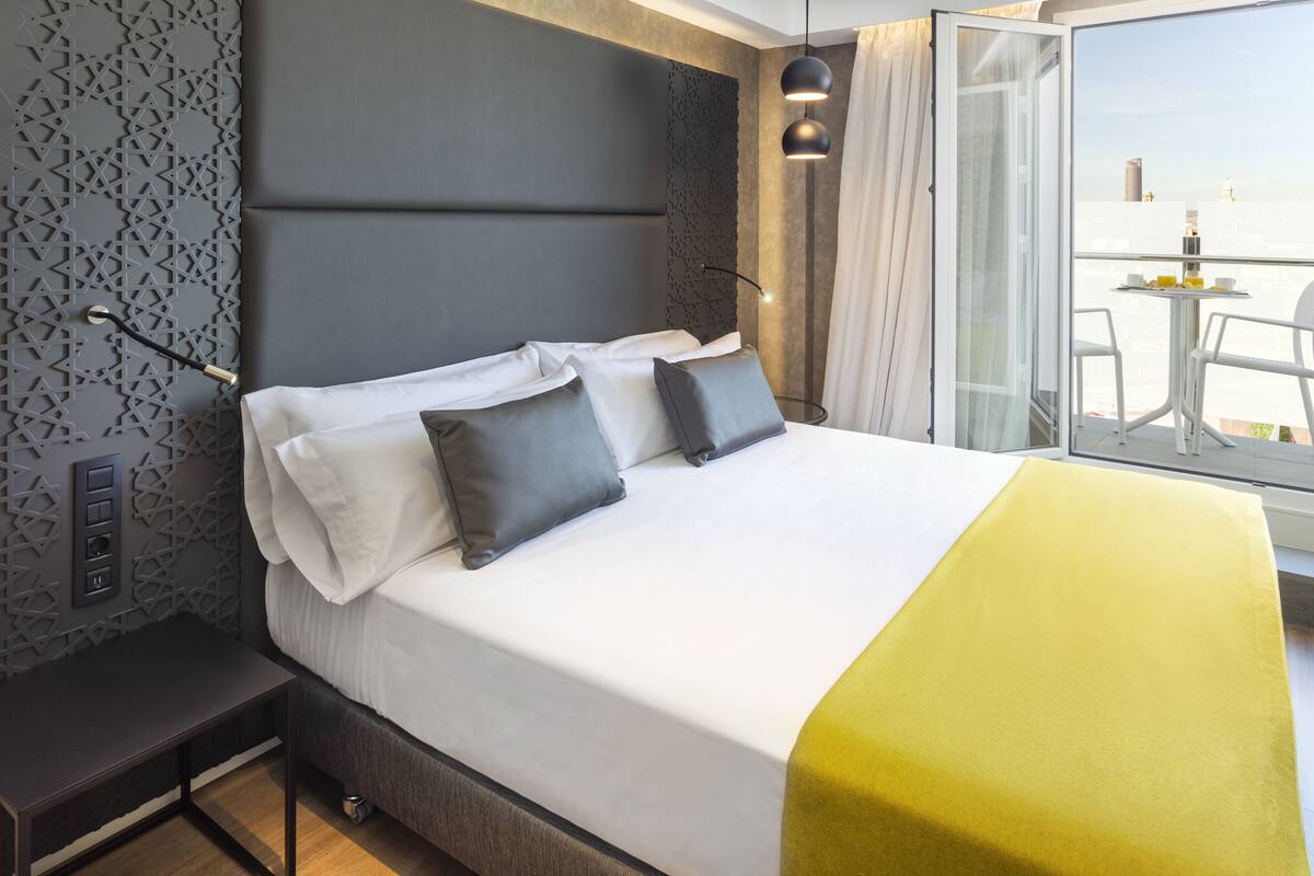 A modern hotel room with a large bed, a bedside table, and a view of the city through a balcony.