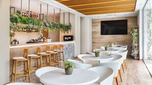 A modern bar area with round tables and wicker chairs, a wine rack with bottles, and a large TV on the wall.