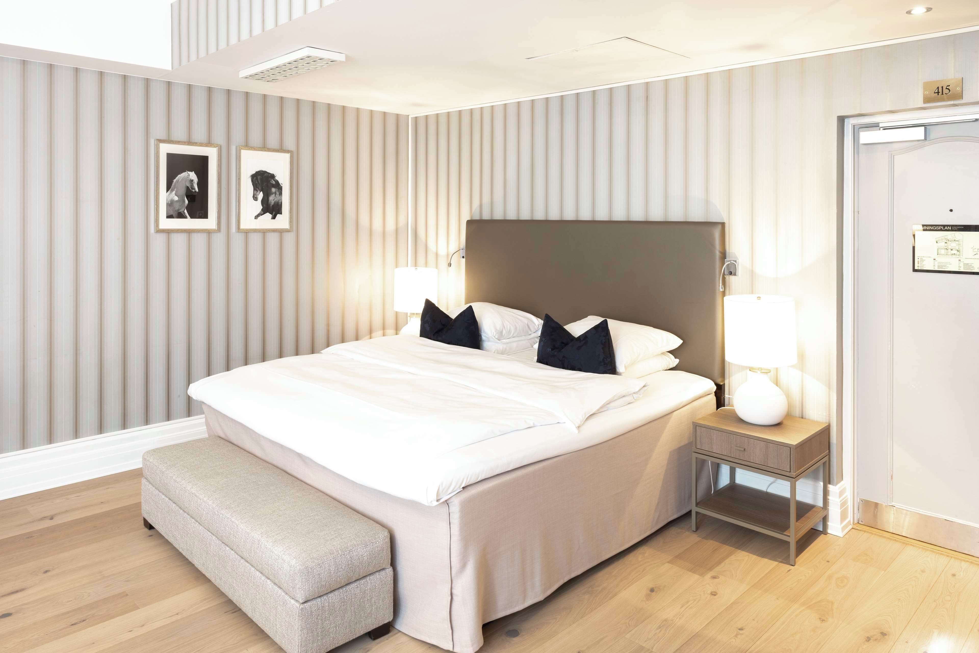 A modern hotel room with a large bed, a bedside table with a lamp, and a bench.