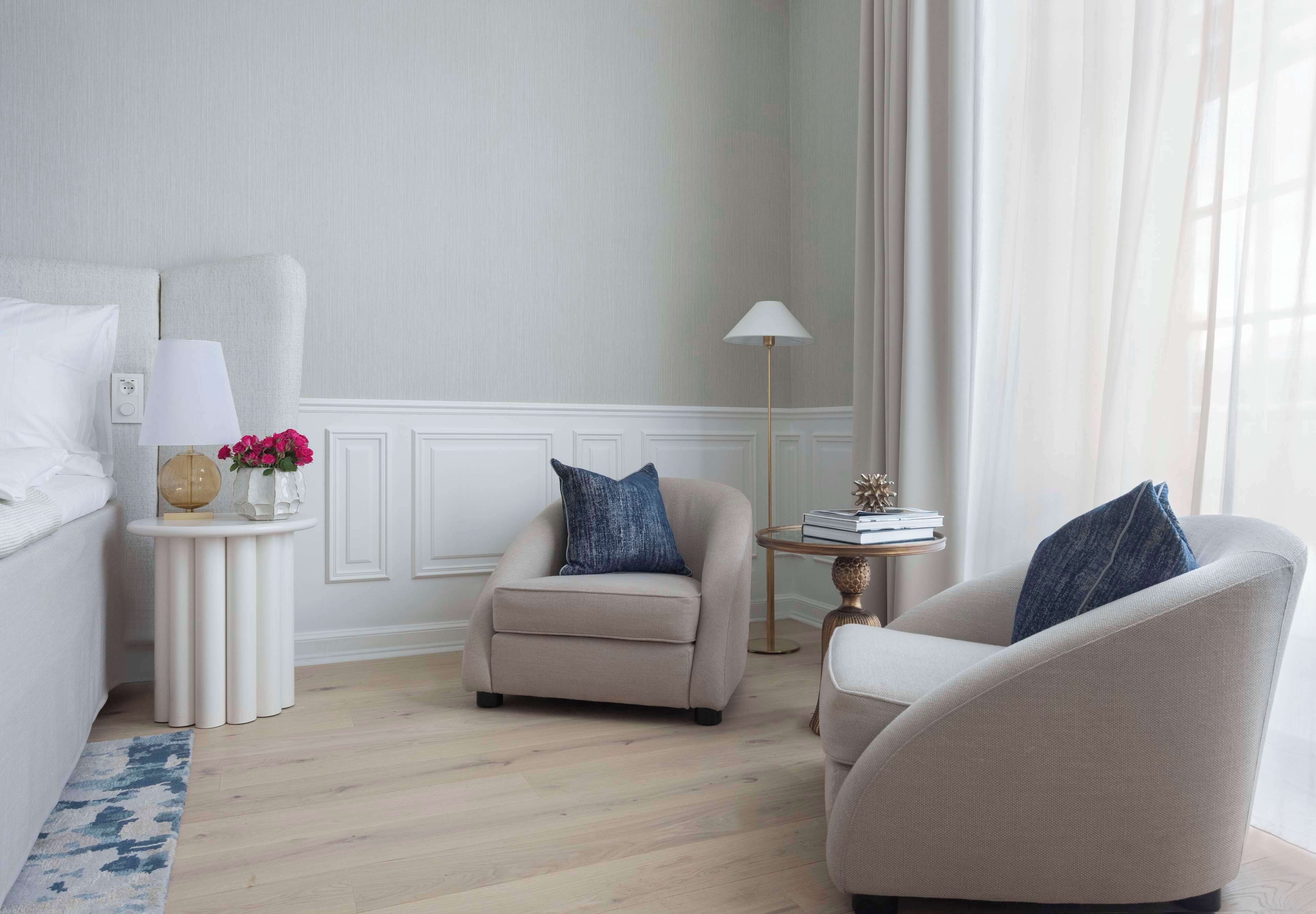 A hotel room with a bed, two armchairs, a small round table with a lamp, and a vase with flowers.