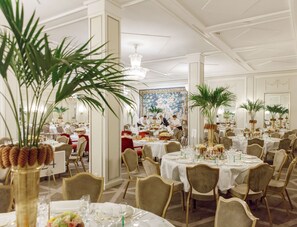A formal dining room with multiple round tables set for guests, each with white tablecloths and gold chairs.