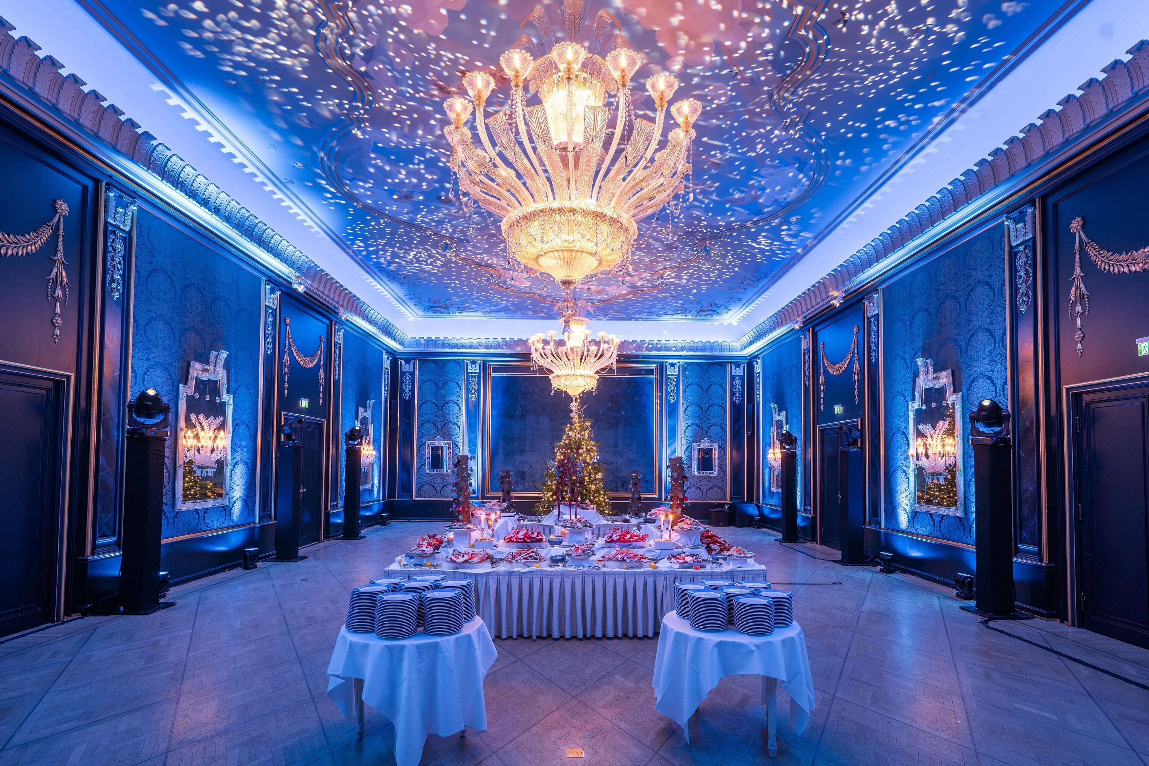 A grand ballroom with a central chandelier, a Christmas tree, and tables set for a formal event.