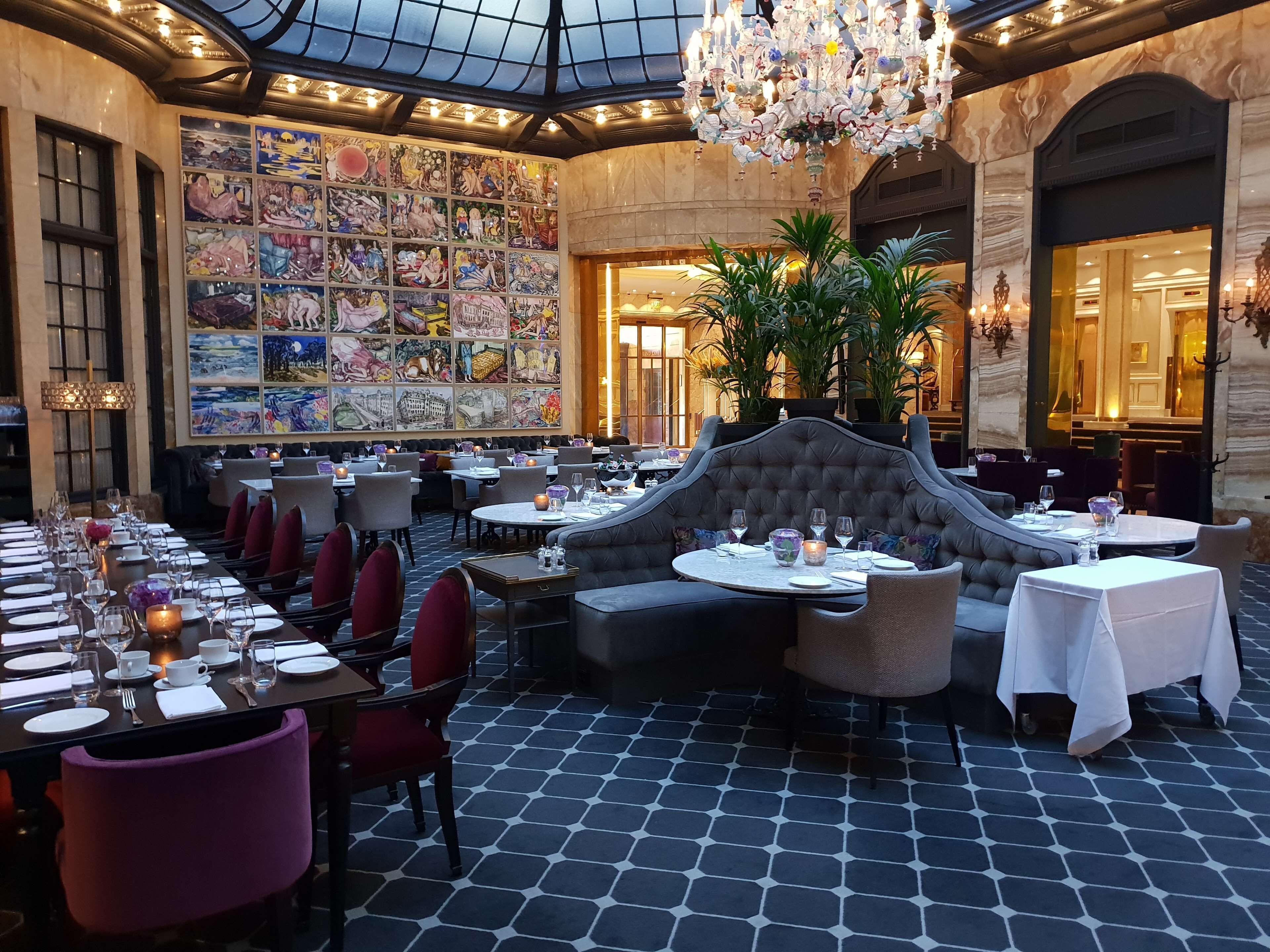 En restaurant med højt til loftet, en lysekrone og en væg med farverige kunstværker. Bordene er dækket med tallerkener, glas og stearinlys.