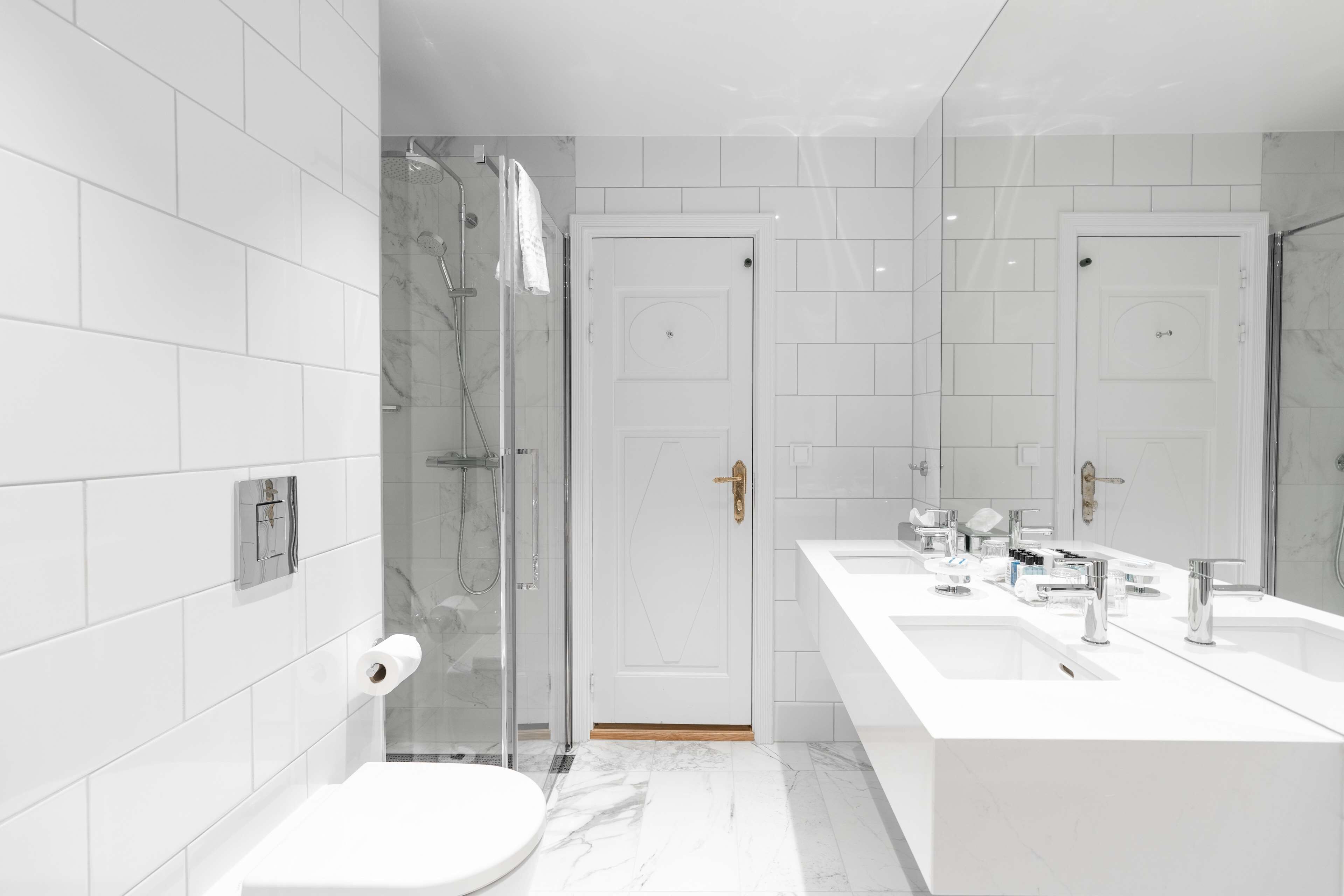 Un baño moderno con baldosas blancas, cabina de ducha de vidrio, inodoro blanco y tocador con dos lavamanos.