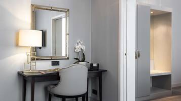 A modern room with a desk, chair, and a large mirror. There is a small table with a lamp and a vase of flowers. A wardrobe is visible in the background.