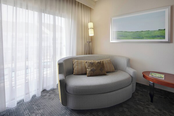 Una habitación moderna con un sofá gris curvo, una pequeña mesita de madera, una lámpara de pie y un cuadro con un paisaje en la pared.