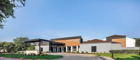 A modern building with a sign reading "Courtyard by Wyndham." It features a covered entrance, a parking lot, and a well-maintained lawn.