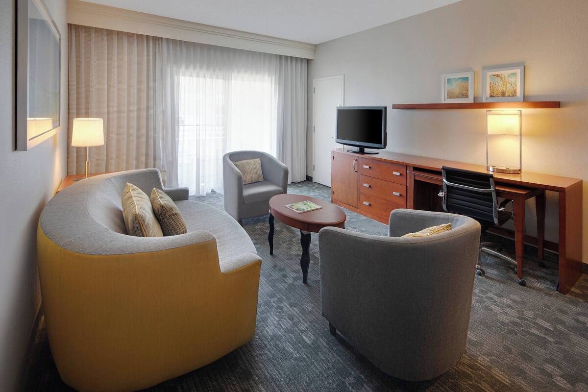 A hotel room with a sofa, armchair, desk, television, and a lamp.