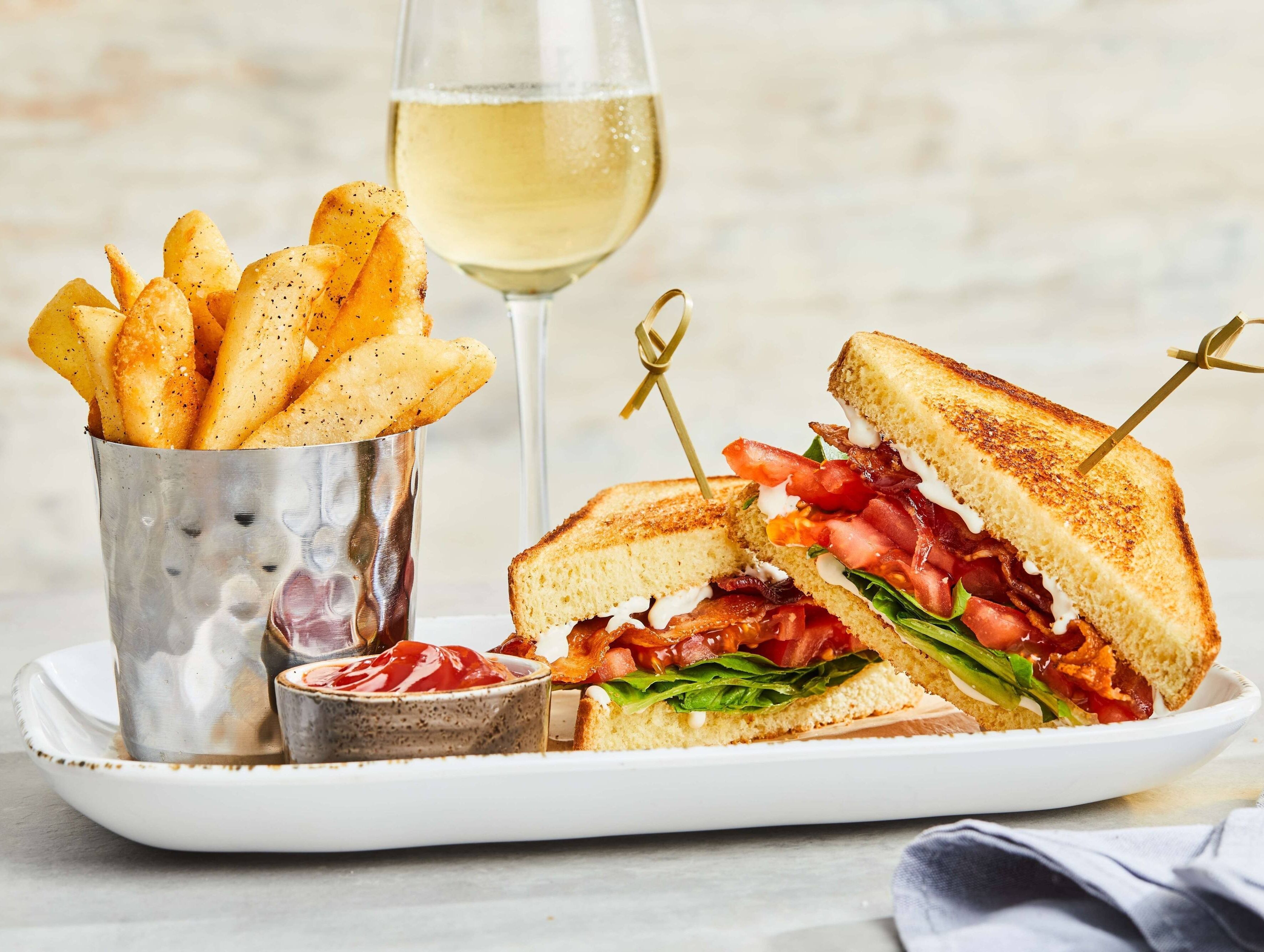 A sandwich cut in half with visible layers of lettuce, tomato, and bacon, accompanied by a glass of white wine and a container of fries.