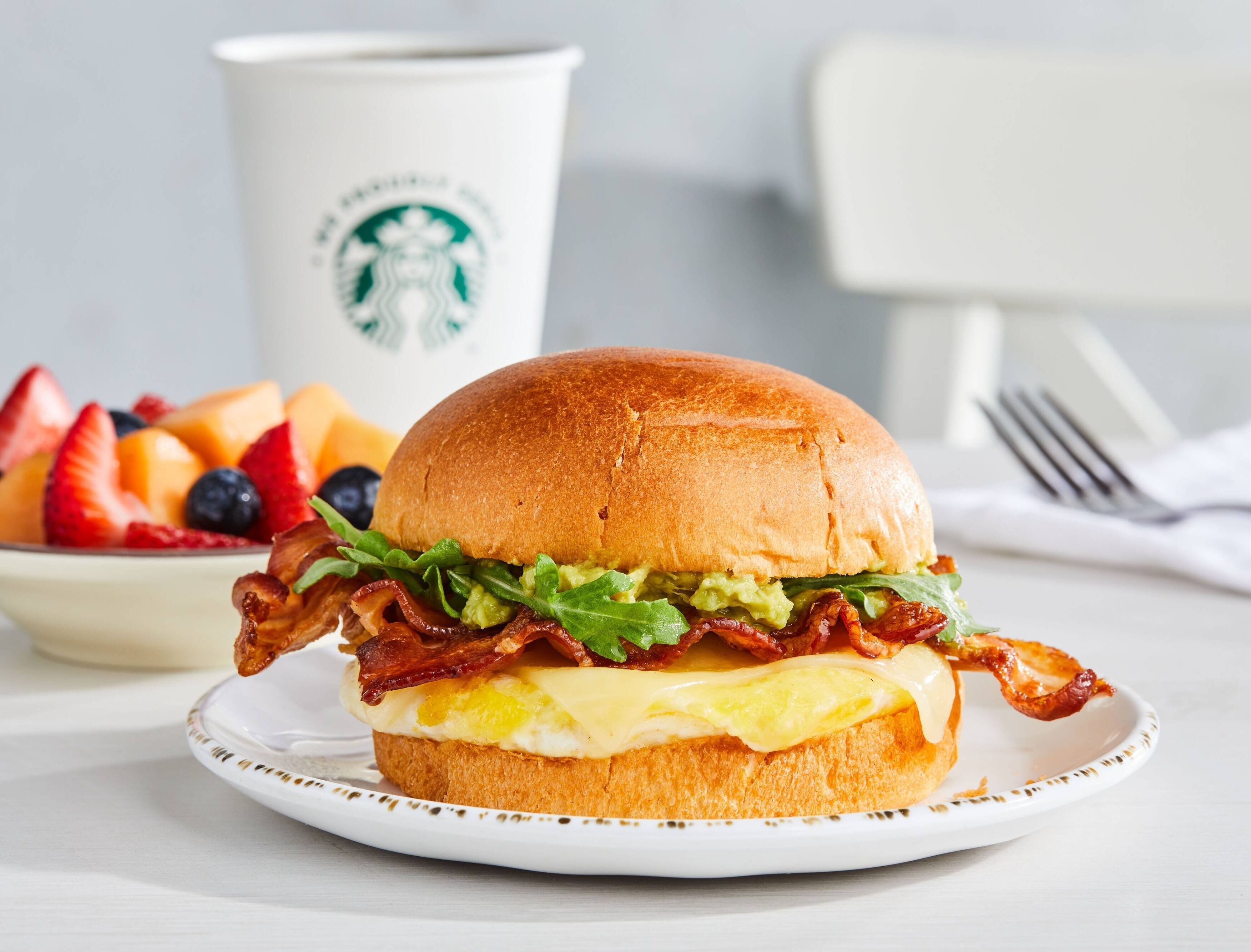 Sanduíche de café da manhã com bacon, alface e ovo no pão, servido em um prato com borda branca.
