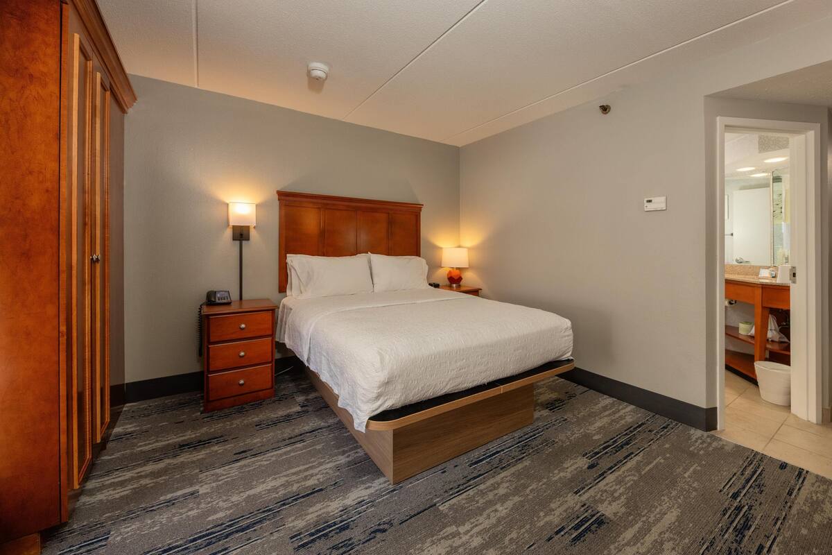 Un dormitorio con cama, lámparas de noche, mesita de noche de madera, armario y baño visible a través de una puerta abierta.