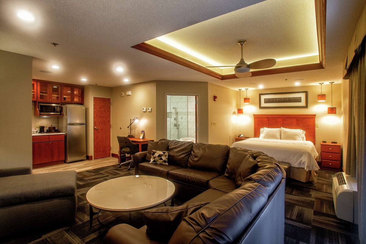 Una habitación de hotel moderna con kitchenette, una zona de estar con sofá y mesa de centro, una cama con cabecera y un ventilador de techo.